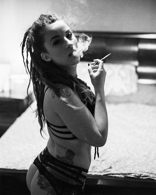 A woman with dreadlocks poses indoors, smoking a cigarette in a black and white artistic shot.