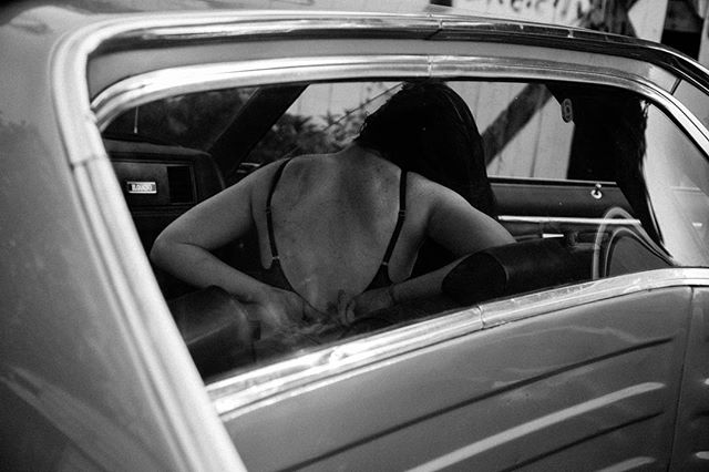 A black and white photo shows a woman in a bra inside a car.