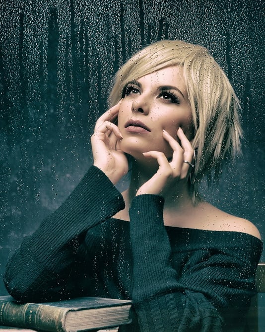 A pensive woman with blonde hair looks up thoughtfully by a rain-streaked window with books.