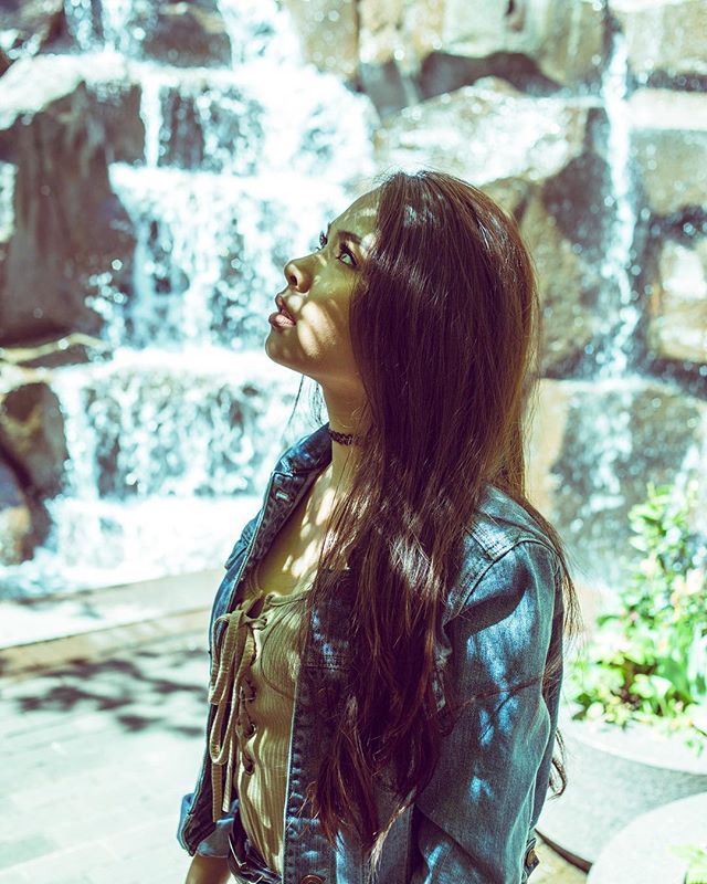 A young woman gazes upward by a waterfall, wearing a denim jacket in a natural outdoor setting.