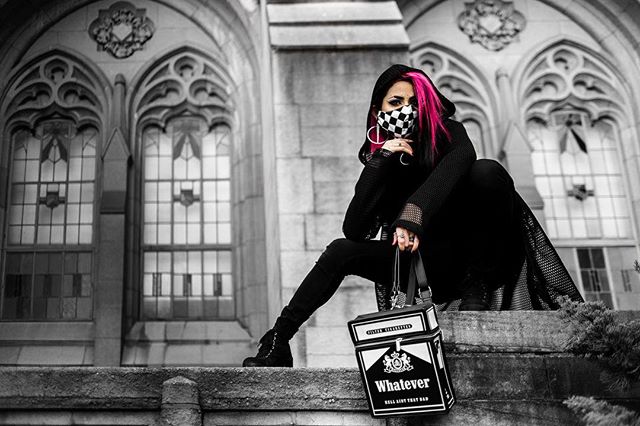 A woman with pink hair sits with a cigarette box bag in front of gothic architecture.