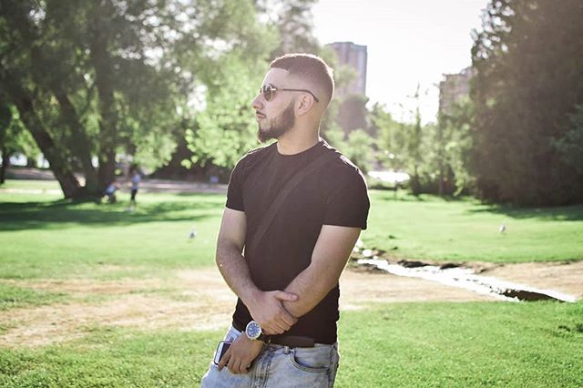 Man with sunglasses in a park, wearing black t-shirt with jeans and holding a phone, looks into the distance.