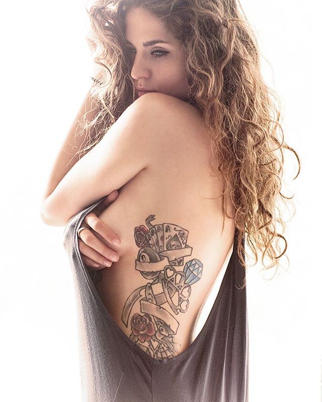 A woman with curly hair poses, showing a detailed tattoo on her back in a softly lit studio.