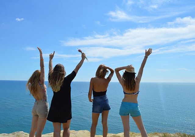 Four young women celebrate by the ocean, enjoying a sunny day together on a scenic coastline.