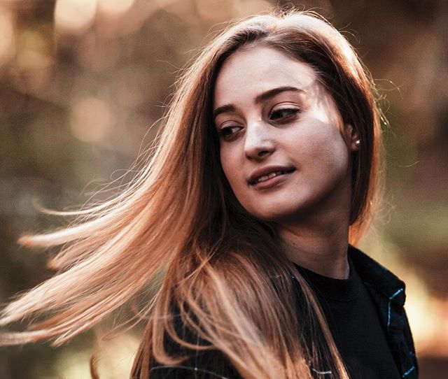 A young woman with long hair thoughtfully looks off into the distance in a serene outdoor setting.