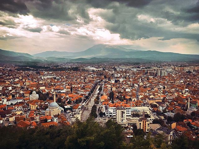 A cityscape with a mosque nestled among buildings, set against a backdrop of mountains under a cloudy sky.