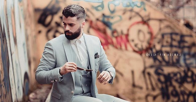 A stylish bearded man in a light gray suit holds sunglasses against a graffiti background.