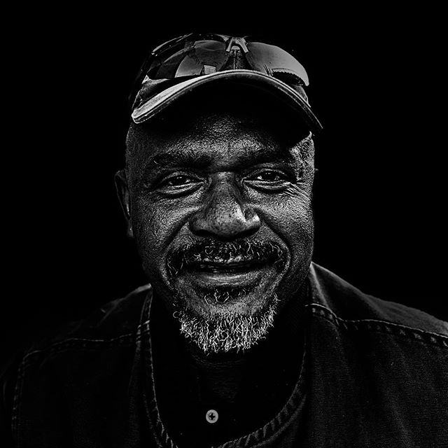 A black and white portrait of a man wearing a cap, captured in a studio against a stark black background.