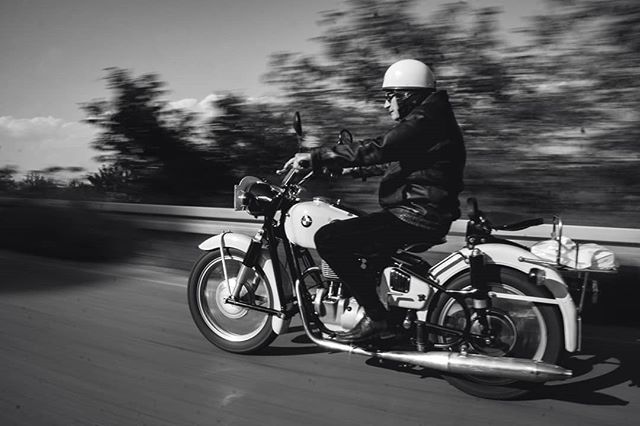 A man wearing a helmet and sunglasses rides a vintage motorcycle on a country road in black and white.
