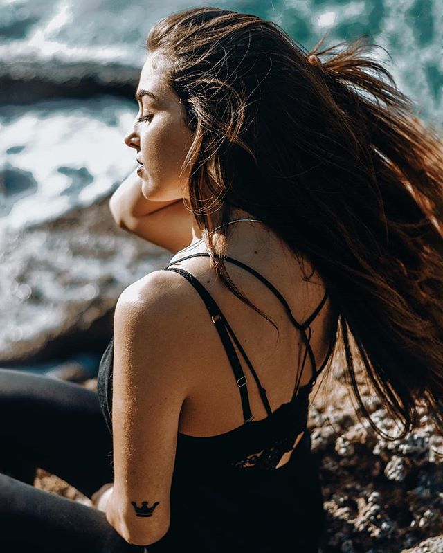 A brunette woman relaxes on the rocky beach with her back to the camera, enjoying the peace.