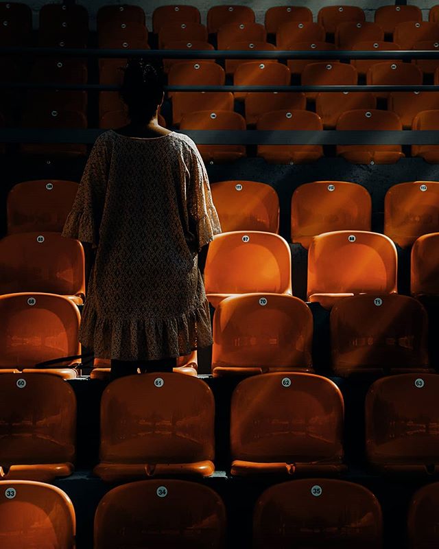 A woman stands alone in a stadium of empty orange seats, creating a sense of solitude and anticipation.