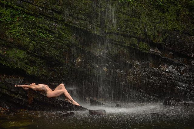 A nude woman reclines on a rock under a waterfall, blending with nature in a serene outdoor setting.