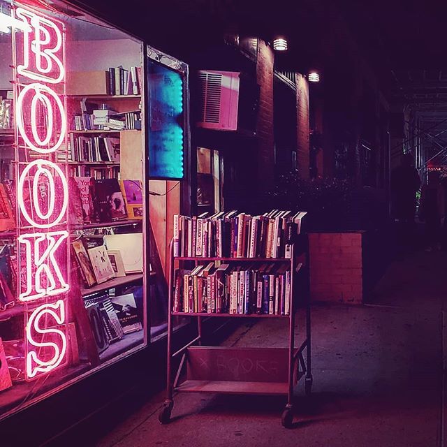 A neon "BOOKS" sign glows next to a street book cart at night, creating an inviting urban scene.