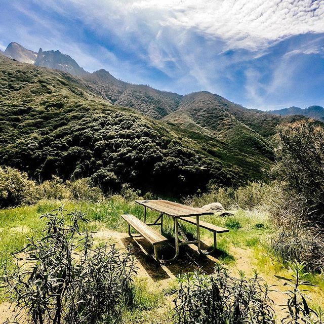 A picnic table sits in a field beneath a mountain range under a cloudy sky.