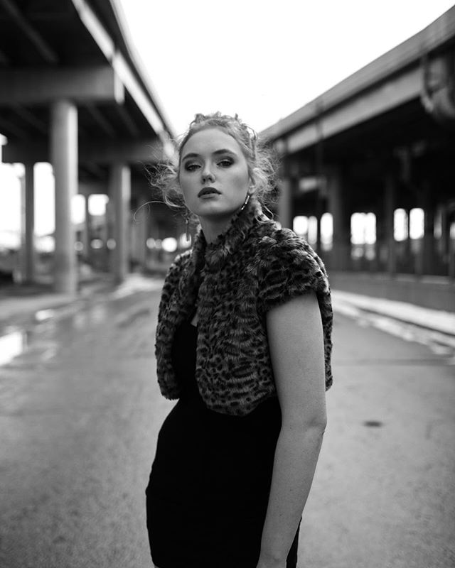 A woman in a black dress and fur shrug poses on a city street in a black and white fashion portrait.
