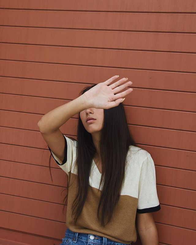 A young woman with long hair poses against a brown wall, shielding her face from the sun with her hand.