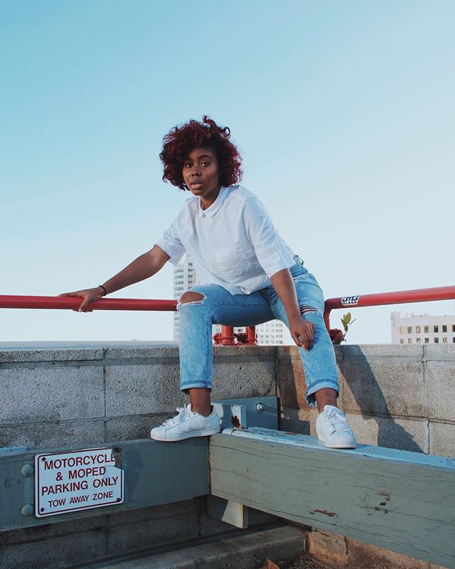 A woman with red hair sits on a ledge in ripped jeans and white sneakers against a blue sky.