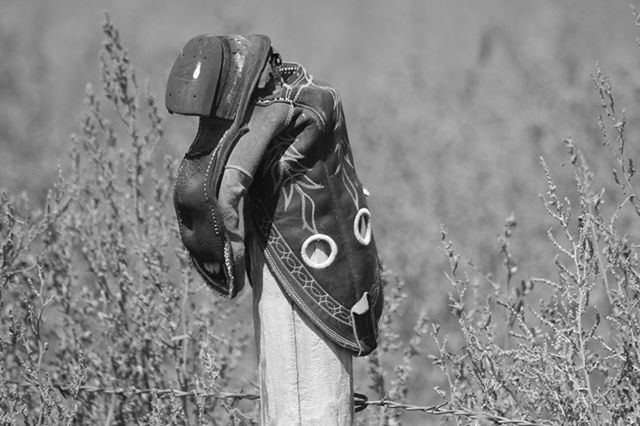 A worn cowboy boot sits atop a weathered fence post in a monochrome rural scene, evoking a sense of nostalgia.