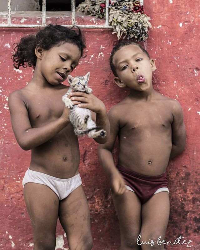 Two young children stand near a red wall, one holding a kitten, capturing a moment of childhood joy.