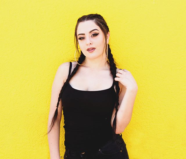 A woman with braids and hoop earrings poses against a yellow wall.