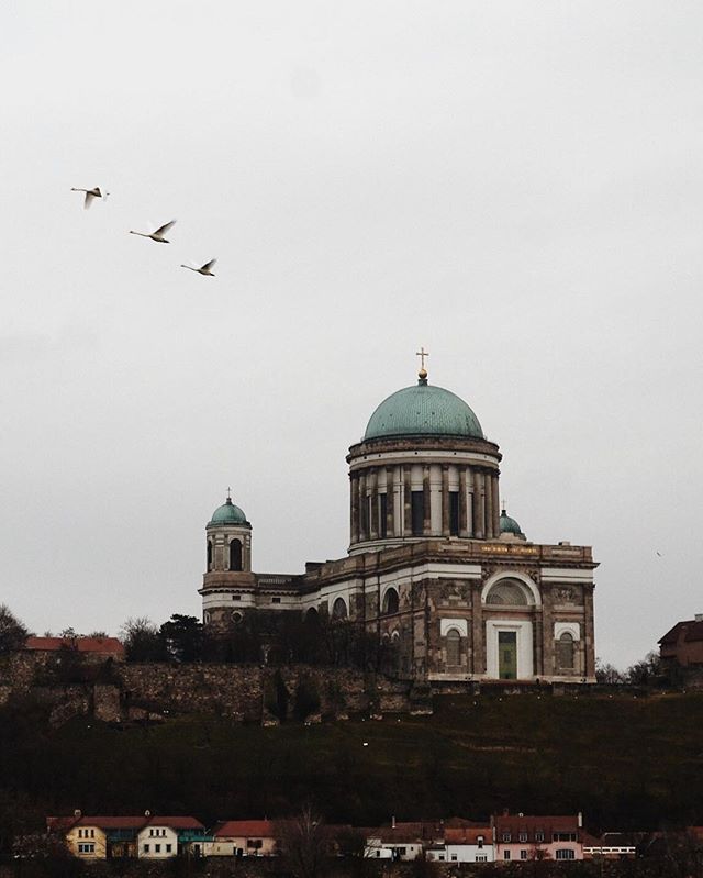 Esztergom Basilica sits atop a hill, with birds flying above, under a cloudy sky.