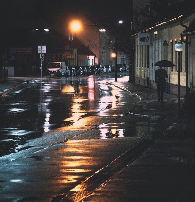 A person with an umbrella walks on a wet city street at night with streetlights reflecting in the puddles.