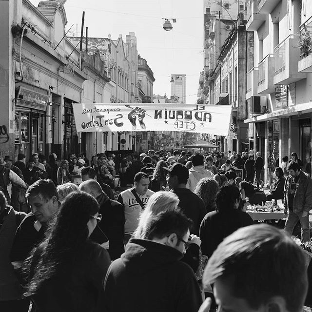 A crowd of people gathers in a city street under a banner during a protest.