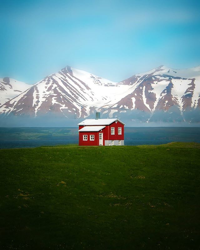 A red house sits in a green field with snowy mountains in the background under a clear sky.