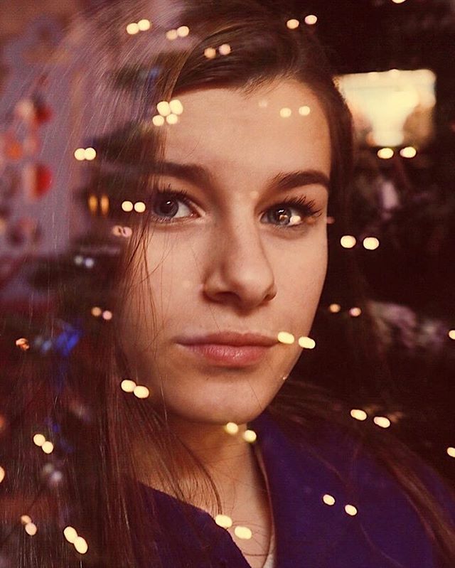 A serene young woman gazes through a window adorned with soft, diffused lights creating a dreamy atmosphere.