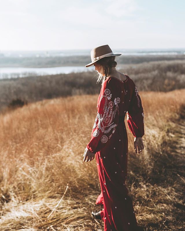 A woman in a red embroidered dress and hat walks through a field on a sunny day.