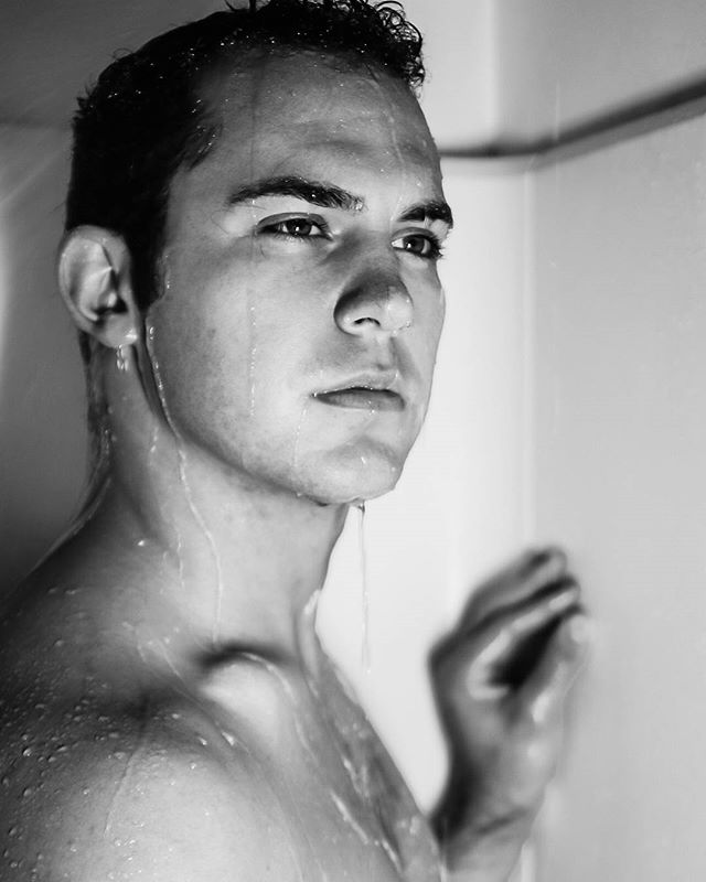 A black and white close-up of a man in the shower with water streaming down his face and body.