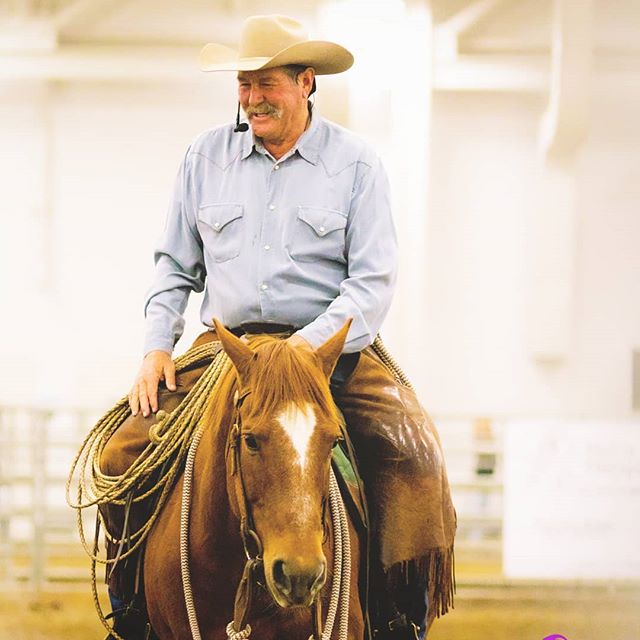 A smiling cowboy wearing a hat rides a horse in an indoor arena.