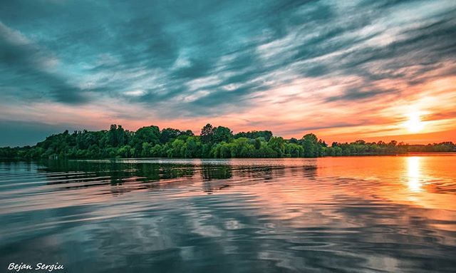 A serene lake reflects the sky at sunset, framed by a green treeline, offering a tranquil nature escape.