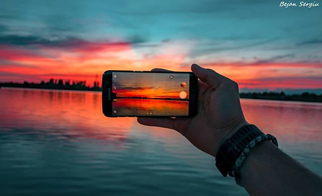 Hand holding a phone taking a picture of a vibrant sunset over the water, capturing the beauty of nature.