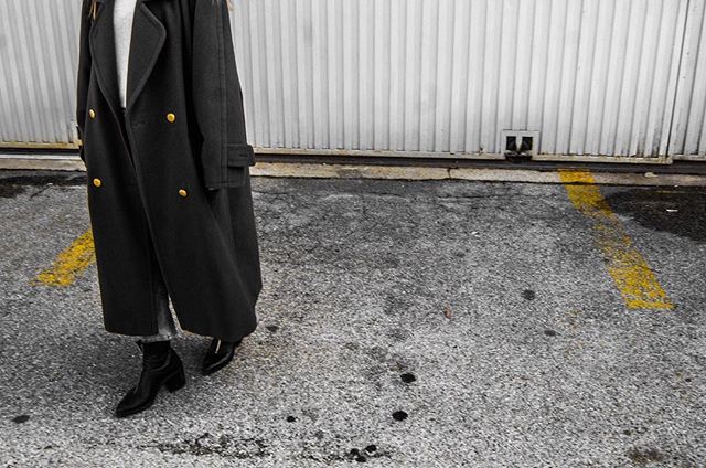 A stylish woman in a long black coat and boots walks along an urban street.