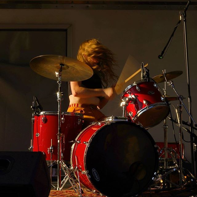 A woman plays a red drum set with cymbals on a stage, performing with energy and passion under natural side lighting.