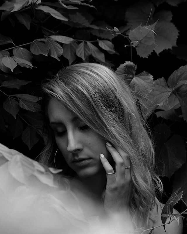 A serene woman surrounded by lush leaves in a dreamy black and white portrait, conveying calmness and intimacy.