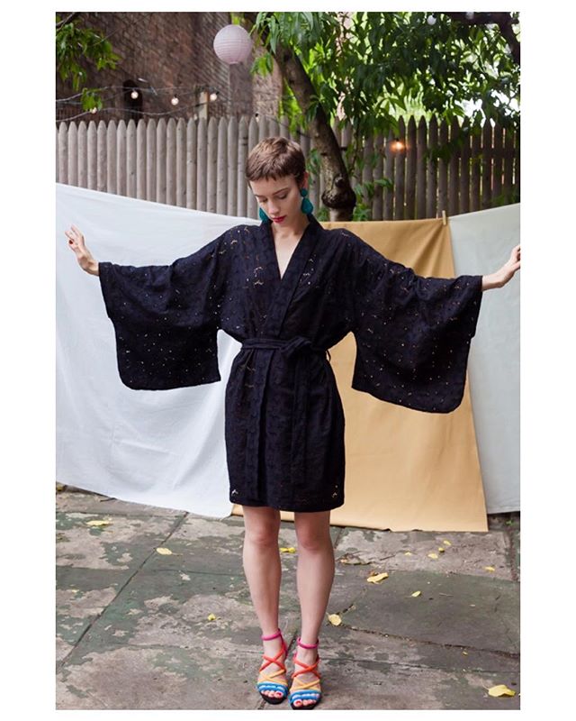 A woman poses in a black kimono dress and colorful sandals in a backyard setting.