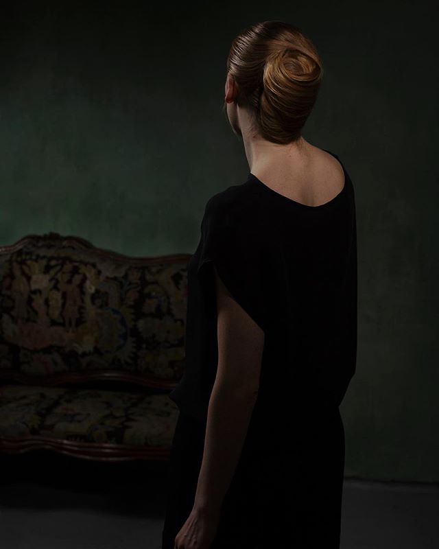 A woman with an elegant updo stands in a studio, wearing a simple black dress, back to the camera.