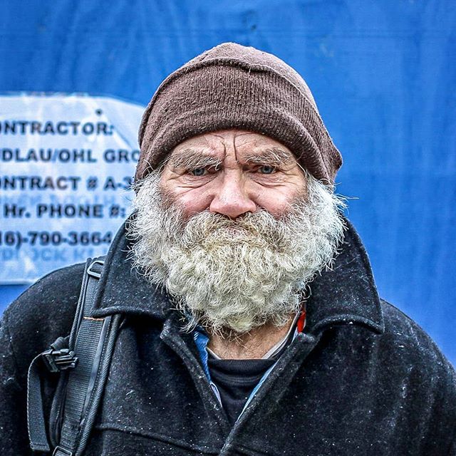 A close-up of an elderly man with a white beard wearing a beanie in an urban setting.
