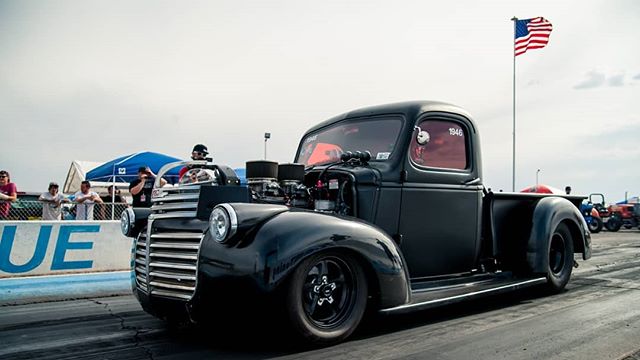 A classic black hot rod truck on a drag strip with an American flag in the background.