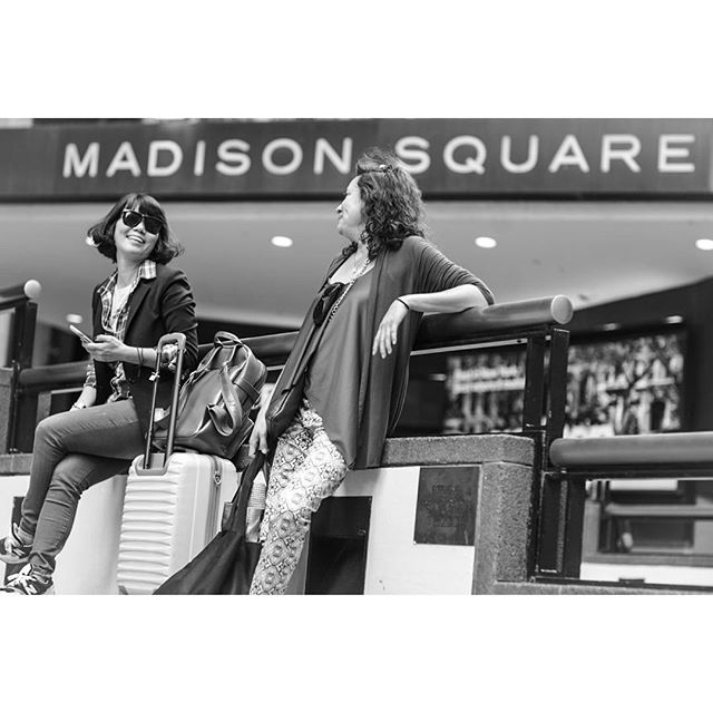 Two women are sitting with their luggage near the Madison Square sign, looking relaxed and chatting.