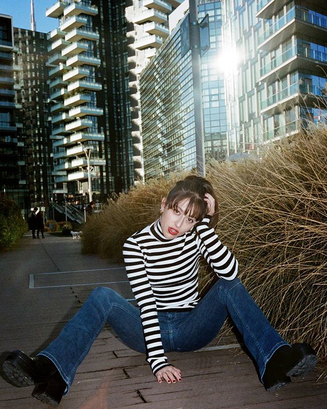 A woman in a striped turtleneck and jeans poses in an urban setting with modern buildings in the background.