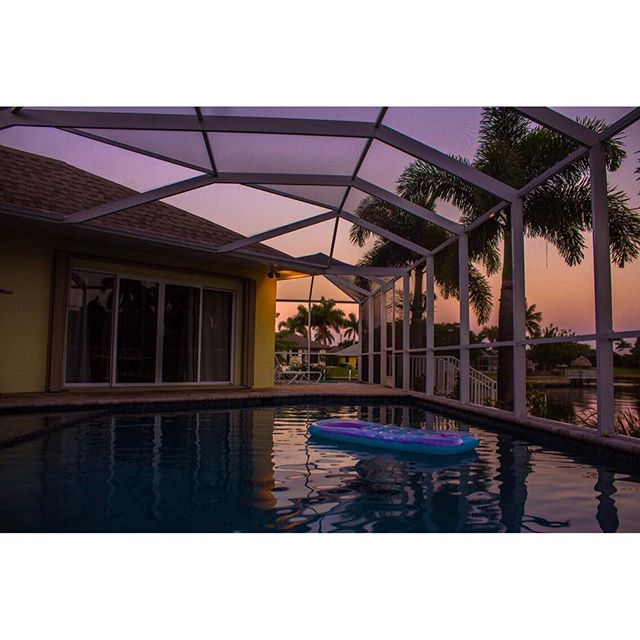 A tranquil pool scene at dusk with an inflatable in the water, palm trees, and a screened-in enclosure.
