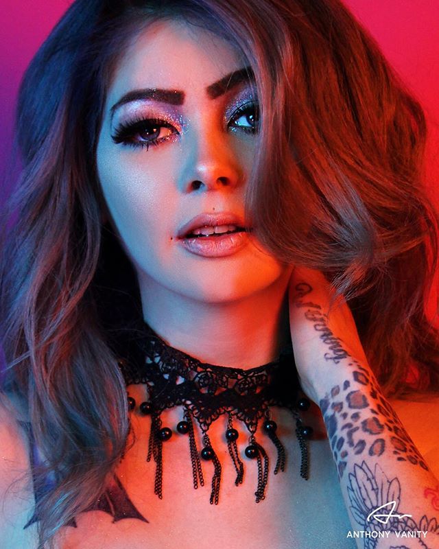 A woman with makeup poses wearing a black lace necklace in a vibrant, colorful studio shot.