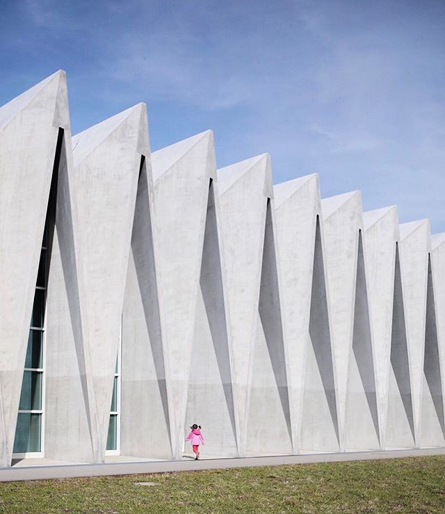 A young girl in a pink coat walks near a modern building with sharp, angular concrete architecture.