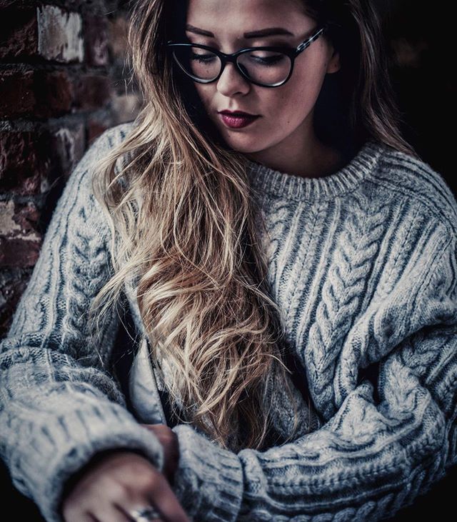 A woman with glasses and a cozy sweater poses against a brick wall in a moody, contemplative portrait.