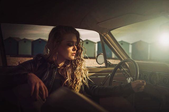 A woman sits in a vintage car, bathed in sunlight, creating a serene and dreamy portrait. 