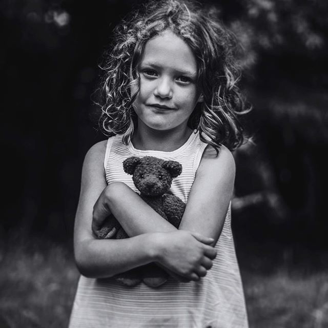 A young girl with curly hair holds a teddy bear in a black and white portrait.