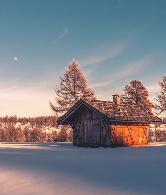 A rustic cabin sits in a snowy landscape under a soft, dreamy sky with a visible moon. 